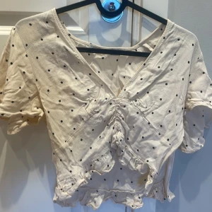 Prickig omlottopp med volangdetaljer - Söt beige omlottopp med svarta prickar och volangdetaljer. Toppen har korta ärmar, v-ringning och rynk framtill. Perfekt för en trendig och feminin look.