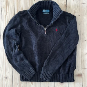 Svart stickad tröja med dragkedja från Polo Ralph Lauren M - Säljer en svart stickad tröja från Polo Ralph Lauren med halv dragkedja och hög krage. Tröjan har ribbade muddar och nederkant samt en liten röd broderad logga på bröstet. Perfekt för kyliga dagar eller bara en laidback stil!