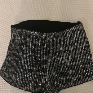 Svarta paljettshorts med dragkedja - Glittriga svarta shorts täckta med silverfärgade paljetter. De har en synlig dragkedja på sidan och en hög midja. Perfekta för att sticka ut och skapa en festlig look.