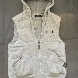 Ralph Lauren Väst - Snygg grå väst med huva från Polo Ralph Lauren. Västen har dragkedja framtill, två fickor och en broderad logga på bröstet. Tillverkad i mjuk bomullsmix med stickad insida.                                    Storlek: S.                                                                           Skick: Mycket bra😊