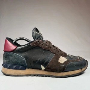 Valentino Rockrunner sneakers i camo och mocka - Säljer ett par Valentino Rockrunner sneakers med camouflagemönster i svart, brunt och beige. Skorna har detaljer i mocka, skinn och en röd häl. Klassisk rund tå och platt sula med nitar på hälen. Perfekta för dig som vill sticka ut med lyxiga detaljer.