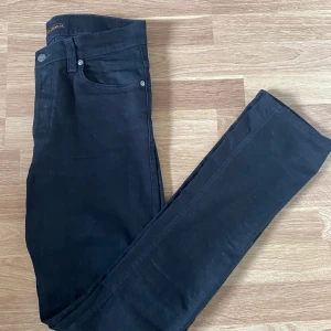 Svarta jeans från Nudie Jeans - Säljer ett par svarta jeans från Nudie Jeans med klassisk femficksdesign och svarta sömmar. Byxorna har en stilren look med Nudie-logga på bakfickan och svart patch i midjan. Perfekta till vardags för en clean stil.