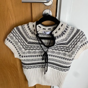 Vit och svart stickad topp från Zara, stl S - Supersöt stickad topp från Zara i vitt med svarta detaljer och mönster, aldrig använd, som ny!Kortärmad modell med rund hals och dekorativ knytning framtill. Perfekt att styla med jeans eller kjol för en trendig look.