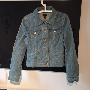 Jeansjacka från H&M - Säljer en tidlös blå jeansjacka från H&M, med guldiga knappar både framtill och på ärmsluten. Jackan har två bröstfickor med lock och två snedställda fickor framtill (de är dekorativa, inte funktionella). En mångsidig jacka som är lätt att kombinera och passar både till vardags och som en stilren detalj i din outfit. Oanvänd, endast prislappen saknas.