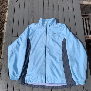 Ljusblå vindjacka från Umbro - Snygg ljusblå vindjacka från Umbro med mörkgrå detaljer på sidorna och ryggen. Jackan har dragkedja framtill, hög krage och meshfoder på insidan för extra komfort. Perfekt för blåsiga dagar och enkel att matcha med olika outfits.