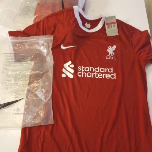 Liverpool FC röd matchtröja från Nike - Säljer en röd Liverpool FC fotbollströja från Nike med vitt tryck och klubbmärke på bröstet. Tröjan har vit krage, korta ärmar och är tillverkad i Dri-FIT-material som andas. Perfekt för fans av LFC!