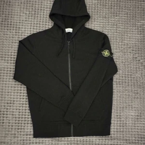 Svart hoodie från Stone Island - Svart hoodie från Stone Island med dragkedja framtill och klassisk logotyp-patch på vänster ärm. Tröjan har huva med dragsko och långa ärmar. Perfekt för en avslappnad och stilren look. Pris kan diskuteras vid snabb affär 