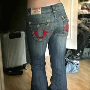 Blå bootcut jeans från True Religion - Säljer ett par blå jeans från True Religion med bootcut passform. Jeansen har röda broderade detaljer på bakfickorna, silverfärgade knappar och klassisk femficksdesign. Perfekta för dig som gillar en retro vibe och markerade detaljer.