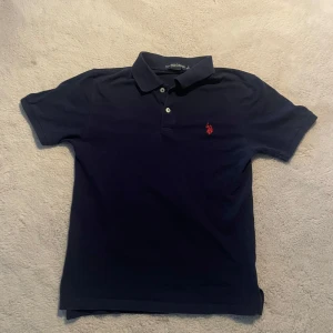 Marinblå pikétröja från U.S. Polo Assn. - Säljer en klassisk marinblå pikétröja från U.S. Polo Assn. i storlek Xs/s. Tröjan har korta ärmar, krage och knappar framtill samt en röd broderad logga på bröstet. Perfekt för varma sommardagar och passar dig som är 158-170