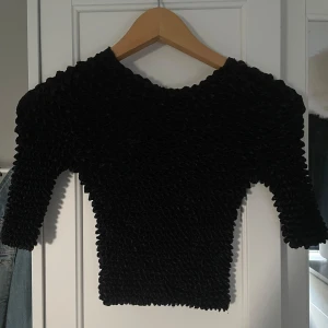 Svart smockad topp med lång ärm - Säljer en svart topp med smockat tyg och lång ärm. Toppen har en tight passform och är tillverkad i ett stretchigt syntetmaterial som ger en cool struktur. Perfekt för dig som vill sticka ut lite extra! Kan bli upp till en M i storlek