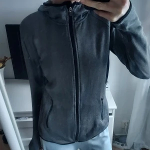 Grå fleecejacka med dragkedja - Mysig grå fleecejacka med dragkedja och huva. Perfekt för kyliga dagar. Jackan har praktiska fickor med dragkedja och långa ärmar.