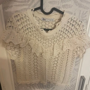 Beige stickad topp med volang från Zara - Säljer en söt beige stickad topp från Zara med korta ärmar och dekorativa volanger i spets över bröstet. Storlek M men sitter som S. Tröjan har ett luftigt, virkat mönster och rund halsringning. Perfekt för vår och sommar!