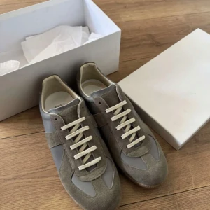 Grå sneakers med mockadetaljer - Säljer ett par grå sneakers från Maison Margiela med snygga mockadetaljer och vita snören. Skorna har en klassisk design med rund tå och platt sula, samt gummisula för bra grepp. Perfekta för dig som gillar stilrena och tidlösa sneakers.