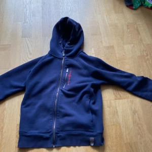 Marinblå hoodie från Sail Racing - Mörkblå hoodie med dragkedja från Sail Racing. Stor logga på ryggen och mindre broderad logga på bröstet i rött. Huva och två fickor framtill. Perfekt för en sportig och avslappnad stil.