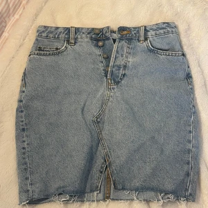 Blå jeanskjol från H&M - Säljer en klassisk blå jeanskjol från H&M med mellan hög midja, knappar framtill och rå kant nertill. Kjolen har fickor både fram och bak samt en liten slits framtill för extra stilpoäng. Helt oanvänd !!