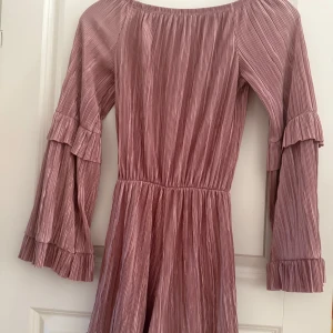 Rosa plisserad jumpsuit med volangärmar - Säljer en superfin rosa jumpsuit med plisserat tyg och vida, långa ärmar med volangdetaljer. Jumpsuit har resår i midjan och en lätt glansig finish. Perfekt för dig som gillar romantisk stil!