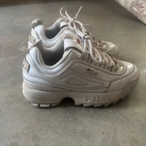 Vita Fila Disruptor sneakers - Säljer ett par klassiska vita Fila Disruptor sneakers med chunky sula och snörning. Skorna har Fila-logga på sidan och sulan, samt detaljer i vitt och lite rött. Perfekta för en sportig och trendig look.
