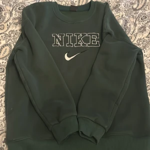 Mörkgrön sweatshirt från Nike - Säljer en mörkgrön sweatshirt från Nike med broderad logga och text på bröstet. Tröjan har rund halsringning och långa ärmar. Perfekt för en avslappnad stil och enkel att matcha med jeans eller mjukisbyxor.
