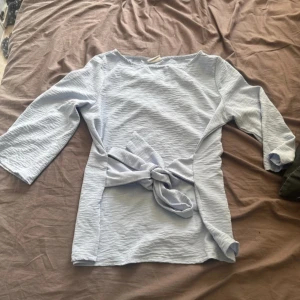 Ljusblå blus med knytband - Säljer en ljusblå blus med trekvartsärm och knytband i midjan. Blusen har en lätt strukturerad yta och rund halsringning. Perfekt för en stilren och enkel look.