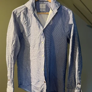 Blåvit mönstrad skjorta från Bläck - Säljer en blåvit långärmad skjorta från Bläck i regular fit. Skjortan har små geometriska mönster, klassisk krage och knappar framtill samt vid ärmslut. Perfekt för dig som gillar stilrena detaljer.