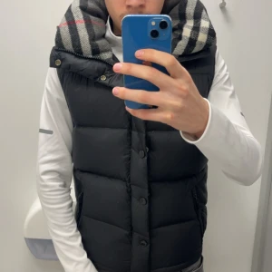 Svart dunväst med rutigt foder - Säljer en svart dunväst med knappar och dragkedja. Västen har ett rutigt foder i huvan med grå, vit och röd färg. Perfekt för lager-på-lager och har en klassisk, puffad look. Jackan kommer ifrån märket Burberry london England. Man kan ta av armarna så att du kan ha på dig den på sommar kvällerna. 