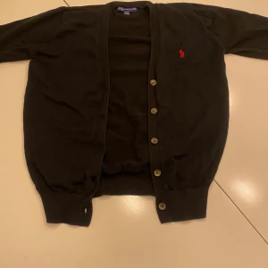 Svart kofta från Ralph Lauren - Svart kofta från Ralph Lauren med klassisk passform och knappar framtill. Tröjan har en liten röd broderad logga på bröstet och är tillverkad i bomull. Perfekt för lager-på-lager och enkel att matcha med olika outfits.