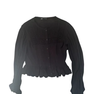 Svart blus med spetsdetaljer från Zara - Säljer en svart blus från Zara med fina spetsdetaljer och knappar framtill. Blusen har lång ärm, volang nertill och broderade blommor på bröstet. Perfekt för dig som gillar romantiska detaljer och en feminin stil.
