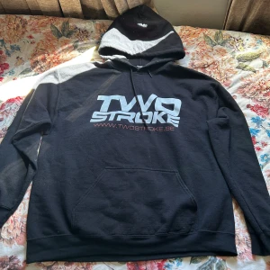Svart hoodie från Twostroke - Säljer en svart hoodie från Twostroke med stor vit logga framtill och färgglad stunt rider-tryck på ryggen. Huva med grå och vit detalj samt magficka. Perfekt för dig som gillar streetwear och motorcyklar.