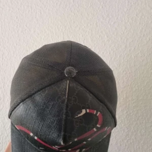 Svart keps med ormtryck från Gucci - Säljer en svart keps från Gucci med ett tydligt ormtryck i rött, vitt och svart framtill. Kepsen har mesh-tyg baktill och justerbar rem. Klassisk Gucci-logga syns diskret i bakgrunden på tyget. Äkta 