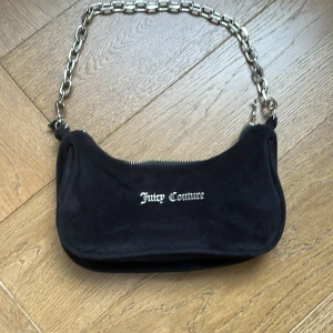 Svart axelväska från Juicy Couture - Säljer en svart axelväska från Juicy Couture med silverfärgad kedjerem och dragkedja. Väskan har en mjuk yta och logga framtill. Perfekt för att lyfta din outfit med en cool detalj. Lite rostig och lite sönder på ena kanten men funkar som ny!❤️⭐️orginal pris:800