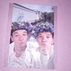 Fotografi med Marcus och Martinus med blomkransar - Säljer ett färgfoto där Marcus och Martinus poserar med blå och gula blomkransar på huvudet. Bilden är tagen utomhus vid ett vattendrag med hus i bakgrunden. Fotot är tryckt på glansigt papper och levereras i plastficka.