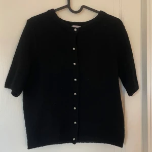 Svart kofta med pärlknappar från H&M - En svart stickad kofta från H&M med korta ärmar och pärlknappar framtill. Enkel och stilren design som passar till många olika outfits. Säljer då den inte kommer till användning 💕