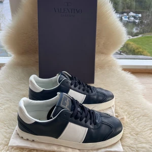 Valentino Flycrews - Säljer ett par snygga Valentino Garavani sneakers i svart och vitt skinn. Skorna har klassisk rund tå, platt sula och coola nitar på hälen. Perfekta för dig som vill ha en stilren men ändå unik look.