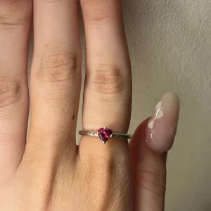 Pandora ring - Säljer en söt silverring från Pandora strlk 16mm tror jag, den har enhjärtformad rosa sten i mitten. Ringen har en tunn, enkel ringskena och små detaljer på sidorna av stenen. Perfekt för dig som gillar romantiska smycken med färg.