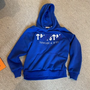 Blå hoodie från Trapstar - Säljer en blå hoodie från Trapstar med vit och blå broderad text på bröstet där det står 'Trapstar' och 'It's a secret'. Tröjan har huva och en stor magficka framtill. Perfekt för dig som gillar streetwear.