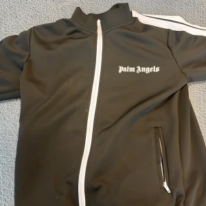 Svart track jacket från Palm Angels - Säljer en svart track jacket från Palm Angels med vit dragkedja och vita ränder längs ärmarna. Jackan har ståkrage, fickor med dragkedja och logga tryckt på bröstet. Perfekt för en sportig och trendig look.