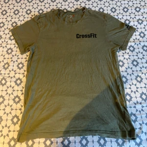 Olivgrön CrossFit t-shirt med tryck - Säljer en olivgrön t-shirt från CrossFit med svart logga framtill och ett stort tryck av en trehövdad hund samt texten 'Mess You Up' på ryggen. T-shirten har rund hals och korta ärmar. Perfekt för sommaren 