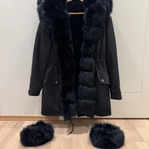 Svart parkajacka med pälskant - Säljer en svart parkajacka med stor huva och lyxig pälskant längs huva och öppning. Jackan har dragsko i midjan, två fickor med lock och knappar framtill. Pälsen är fluffig och ger en exklusiv känsla. Perfekt för kyliga dagar. Jackan har äkta päls över hela!!! Storlek M!!! Priset kan diskuteras!! 🥰🥰🥰🥰 Nypris: 4000kr