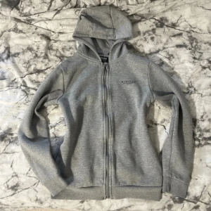 Grå hoodie från McKenzie - Säljer en klassisk grå hoodie från McKenzie med dragkedja framtill och huva. Tröjan har diskret logga på bröstet och är tillverkad i mjukt bomullsmaterial. Perfekt för en avslappnad stil.