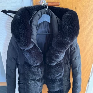 Svart dunjacka med fuskpäls - Säljer en svart dunjacka med stor, fluffig fuskpäls runt kragen och längs dragkedjan. Jackan har en pufferstil och ger en lyxig känsla med sin breda pälsdetalj. Perfekt för kalla dagar när du vill hålla dig varm och snygg.
