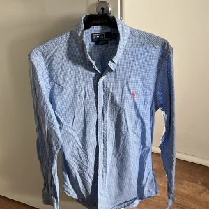 Blå- och vitrutigt skjorta från Polo Ralph Lauren - Snygg blå- och vitrutigt skjorta från Polo Ralph Lauren med klassisk krage och rosa broderad logga på bröstet. Slim fit-modell med långa ärmar, perfekt för en stilren look. Lite skrynklig på bilden men annars inga defekter 9/10 skick!