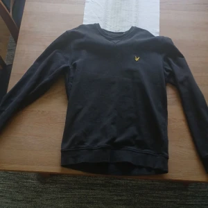 Svart v-ringad tröja från Lyle & Scott - Svart v-ringad tröja från Lyle & Scott med den klassiska gula loggan broderad på bröstet. Tröjan har långa ärmar och är perfekt för en stilren och enkel look.