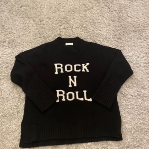 Svart stickad tröja med texten 'Rock N Roll' - Cool svart stickad tröja med vit text 'Rock N Roll' framtill. Tröjan har långa ärmar och ribbade muddar vid ärmslut och nederkant. Perfekt för dig som gillar en avslappnad och trendig stil.