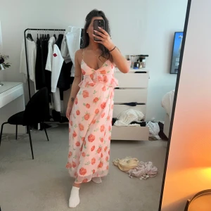 Blommig långklänning med volang - Säljer en söt långklänning med rosa och röda blommor på vit botten. Klänningen har smala axelband, v-ringning och volangdetalj över bysten. Perfekt för sommarens alla tillfällen! Använd 1 gång