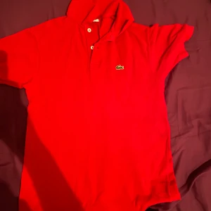 Röd pikétröja från Lacoste - Säljer en klassisk röd pikétröja från Lacoste med den ikoniska krokodilloggan på bröstet. Tröjan har korta ärmar och knappar vid kragen. Perfekt för en stilren och sportig look.