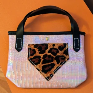 Iridescent handväska med leoparddetalj - Snygg handväska med iridescent yta som skiftar i rosa och blått. Väskan har svarta handtag och en cool leopardmönstrad ficka framtill. Perfekt för att ge din outfit en unik touch!