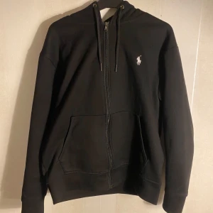 Svart hoodie från Ralph Lauren - Säljer en stilren svart hoodie från Ralph Lauren med dragkedja och det klassiska logotypbroderiet på bröstet. Tröjan har långa ärmar och en praktisk känguruficka framtill. Perfekt för en avslappnad look.