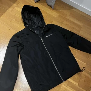  vindjacka Balmain Paris jacka  - Snygg svart vindjacka från Balmain med dragkedja och huva. Jackan har Balmain-logga både fram och bak, vilket ger en stilren look. Perfekt för blåsiga dagar.