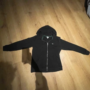 Zip hoodie från Lacoste - Snygg svart hoodie från Lacoste med dragkedja och huva. Den har en liten logga på bröstet och gröna detaljer vid huvan. Perfekt för en avslappnad stil. Bara skriv om ni undrar något.