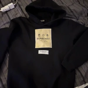 Svart hoodie från Burberry - Säljer en stilren svart hoodie från Burberry med en stor beige logga på bröstet. Hoodien har en klassisk design med huva och långa ärmar. Perfekt för en avslappnad och trendig look.
